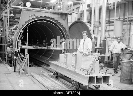 Marc Smits metal gluer Fokker, Whizgle News, Dutch Desk, Paesi Bassi, 1950 - 2000 il 08-11-1990. Questi argomenti sono mostrati nell'immagine. In un ambiente industriale spazioso, una grande camera circolare domina lo sfondo, suggerendo un ambiente di test o produzione. La superficie metallica grigio scuro della camera contrasta con la luminosità dell'area circostante, riflettendo un'area di lavoro ben mantenuta. In primo piano, un uomo si trova con sicurezza accanto a una piattaforma gommata che trasporta diversi pezzi di metallo rettangolari, progettati per complesse attività ingegneristiche. Lui è vestito in un l Foto Stock
