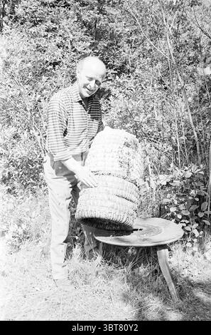 Apicoltore con alveare, Whizgle News, Dutch Desk, Paesi Bassi, 1950 - 2000. L'immagine contiene questi argomenti. Un uomo si trova accanto ad un piccolo tavolo rotondo in legno in un ambiente naturale all'aperto. La sua espressione è calda e amichevole, accompagnata da un leggero sorriso mentre si impegna con lo spettatore. Indossa una camicia button-up a righe, che contrasta con i suoi pantaloni di colore chiaro. Nelle sue mani, bilancia con cura un grande cesto intrecciato, mostrando i suoi intricati motivi e la sua maestria artigianale. L'ambiente circostante è rigoglioso e verde, pieno di fitte foglie e di una varietà di piante che creano Foto Stock