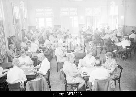 Gli anziani hanno la serata della carta., Whizgle News, Dutch Desk, Paesi Bassi, 1950 - 2000. L'immagine include questi argomenti. La scena è ambientata in una sala spaziosa e animata piena di numerosi tavoli rotondi, ognuno circondato da gruppi di persone impegnati in conversazioni e giochi di carte. La tavolozza dei colori generale è attenuata, caratterizzata prevalentemente da varie sfumature di grigio, che donano un'atmosfera nostalgica. La sala è illuminata da una luce naturale diffusa che scorre attraverso le grandi finestre, creando una luce soffusa che mette in risalto i volti dei partecipanti e proietta ombre delicate sul pavimento. T Foto Stock