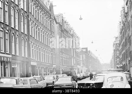 Street scene Amsterdam., Whizgle News, Dutch Desk, Paesi Bassi, 1950 - 2000. Questi argomenti sono mostrati nell'immagine. La scena cattura una vivace strada urbana fiancheggiata da eleganti edifici in mattoni a più piani che presentano grandi finestre con modanature decorative. Queste strutture incarnano uno stile architettonico classico, caratterizzato da un pattern ripetitivo che trasmette un senso di ordine. La tavolozza dei colori è sfumata, dominata da sfumature di grigio e marrone, che trasmettono un'atmosfera vintage. La strada è piena di una varietà di auto parcheggiate di epoche diverse, mostrando una varietà di forme e dimensioni. Il Foto Stock