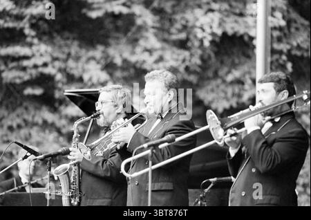 Uralski Jazz Band B'daal Bloemendaal, Whizgle News, Dutch Desk, Paesi Bassi, 1950 - 2000 il 06-07-1994. L'immagine include questi argomenti. In un vivace ambiente all'aperto, tre musicisti suonano strumenti di ottoni, trasudando un'aura di cameratismo e arte. La scena è dominata da tonalità ricche e monocromatiche che creano un senso di nostalgia. Il primo musicista, a sinistra, ha un sassofono, la sua espressione facciale è concentrata e appassionata mentre si impegna con la musica. Indossa occhiali sportivi e ha una barba ben curata, sottolineando il suo comportamento serio. Il secondo musicista occupa il centro, Foto Stock