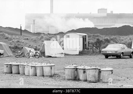 Wijk aan Zee 100 anni di località balneare nel fumo di Hoogovens Blastovens Overviews Wijk aan Zee, Whizgle News, Dutch Desk, Paesi Bassi, 1950 - 2000 il 14-07-1980. Questi sono gli elementi dell'immagine. La scena presenta un ambiente esterno robusto, caratterizzato da un mix di tende e varie strutture temporanee. Un piccolo rimorchio bianco si trova in posizione prominente al centro, circondato da una dispersione di tende di tela che suggeriscono un campeggio improvvisato. Il terreno è irregolare e sembra essere un mix di sporco e ghiaia, punteggiato da diversi grandi contenitori metallici allineati in primo piano, perh Foto Stock