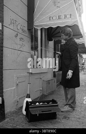 Man rimuove graffiti dal muro di un negozio del centro, Whizgle News, Dutch Desk, Paesi Bassi, 1950 - 2000 il 01-12-1980. L'immagine contiene questi argomenti. In una scena di strada intrisa di un senso di nostalgia, un uomo di mezza età si trova di fronte a un muro di mattoni segnato da vari simboli e graffi, suggerendo una lunga storia di interazioni da parte dei passanti. Vestito con un cappotto scuro e sartoriale, è posizionato leggermente a sinistra, conferendogli una presenza che contrasta con la facciata testurizzata dell'edificio alle sue spalle. La sua postura è dinamica; si inclina leggermente in avanti, manipolando un lungo, Foto Stock