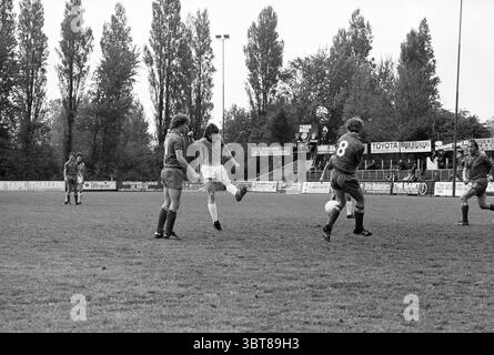 Partita di calcio EDO, Whizgle News, Dutch Desk, Paesi Bassi, 1950 - 2000 il 25-05-1979. L'immagine contiene questi argomenti. In un campo erboso segnato per il calcio, una scena vivace si svolge durante una partita. L'ambiente è un'arena sportiva aperta circondata da alti alberi che aggiungono un tocco di natura a un cielo coperto, creando uno sfondo silenzioso. In primo piano, un giocatore con una maglia bianca viene catturato a metà azione, sollevando una gamba mentre colpisce la palla, mostrando un momento di atletismo e concentrazione. Nelle vicinanze, due giocatori con maglie blu sono posizionati in modo difensivo, ognuno con posizioni diverse: Una b Foto Stock
