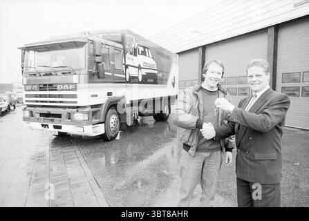 Nuova macchina didattica da tempo Baars, Whizgle News, Dutch Desk, Paesi Bassi, 1950 - 2000 il 01-11-1990. L'immagine contiene questi argomenti. La scena cattura un momento di festa e cameratismo fuori da un grande edificio industriale, probabilmente una concessionaria o un magazzino. In primo piano, due uomini sono impegnati in una stretta di mano, simboleggiando una transazione di successo. Un uomo, vestito con una giacca di pelle scura, appare rilassato e casual, mentre l'altro, con un abito e una cravatta, trasmette professionalità. Dietro di loro si trova un grande veicolo DAF, posizionato in modo preminente, che mostra il suo design audace con un sorprendente Foto Stock