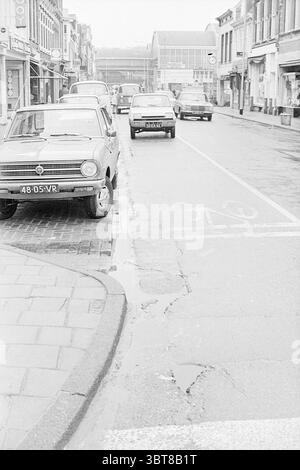 Cars in a shopping Street Haarlem Kruisweg Paesi Bassi., Whizgle News, Dutch Desk, Paesi Bassi, 1950 - 2000. L'immagine mostra questi argomenti. La scena raffigura una tranquilla strada urbana, caratterizzata dalle sue tonalità di grigio e dall'atmosfera ricoperta. Da un lato, una serie di negozi modesti con vetrine espone una varietà di merci, anche se le specifiche non sono distinguibili. Gli edifici sono leggermente usurati, indicativi dell'età, con caratteristiche architettoniche come piccole tende da sole e finestre. In primo piano, diversi veicoli occupano la strada, parcheggiati lungo il marciapiede. Un'auto d'epoca si distingue per la sua Foto Stock
