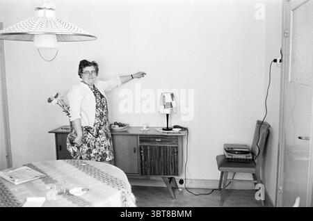 Poltergeist nell'edificio della coppia Kloos Spanjevaartstraat 9 Haarlem Spaansevaartstraat Paesi Bassi, Whizgle News, Dutch Desk, Paesi Bassi, 1950 - 2000 il 06-1961. Questi sono gli argomenti nell'immagine. In una sala illuminata con luce soffusa, una donna si trova in una moderna credenza della metà del secolo. Ha i capelli scuri e corti e indossa occhiali, trasudando un'aria di autorità e calore. Il suo outfit è composto da un abito a motivi geometrici, completato da un cardigan di colore chiaro, che contrasta piacevolmente con le pareti dalle tonalità neutre. La credenza, che presenta texture di legno naturale, è adornata con vari obj Foto Stock
