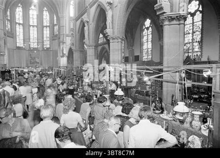 Fiera dell'antiquariato in una chiesa., Whizgle News, Dutch Desk, Paesi Bassi, 1950 - 2000. L'immagine contiene questi argomenti. L'ambiente e' un ampio interno di una grande struttura, probabilmente un edificio storico, date le caratteristiche architettoniche quali alti soffitti a volta adornati da disegni intricati. L'atmosfera è piena di attività, piena di persone impegnate in varie interazioni. L'atmosfera è vivace ma leggermente caotica, sottolineata da un senso di comunità ed eccitazione. La luce si riversa attraverso le finestre ornate di vetro colorato, gettando motivi colorati sul pavimento e. Foto Stock