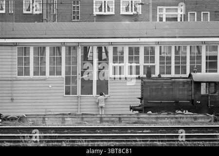 Cape train presso i treni officina NS, Whizgle News, Dutch Desk, Paesi Bassi, 1950 - 2000 il 22-06-1977. L'immagine contiene questi argomenti. La scena cattura un'atmosfera vintage, caratterizzata da toni tenui e un senso di nostalgia. In primo piano, una figura solitaria è vista vicino ad una struttura in legno, che ha una serie di grandi finestre rettangolari. La persona sembra essere impegnata in un'attività vicino a una delle finestre aperte, suggerendo curiosità o forse la necessità di comunicare con qualcuno all'interno. A destra, una vecchia locomotiva a vapore è silenziosa, con la sua superficie scura e ruvida che contrasta Foto Stock