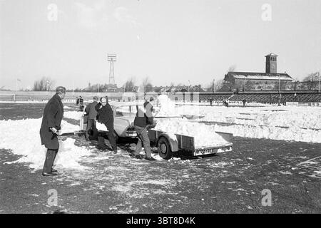 Disinnevamento in Haarlem Grounds Football Haarlem Work, Whizgle News, Dutch Desk, Paesi Bassi, 1950 - 2000 il 04-02-1968. Questi sono gli elementi dell'immagine. La scena cattura un paesaggio invernale, dominato da un grande campo aperto coperto da uno spesso strato di neve. Il cielo sopra è un grigio pallido e coperto, che suggerisce un'atmosfera fredda. In primo piano, diversi individui sono attivamente impegnati nella rimozione della neve, creando macchie chiare sul campo e i loro respiri visibili nell'aria ghiacciata. I lavoratori sono vestiti con cappotti e cappelli scuri, in contrasto con la neve bianca brillante. Qualche ar Foto Stock
