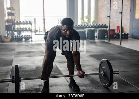 Atleta afroamericano che indossa abiti da palestra sollevando barcampanella carica in palestra con scaffali stupidi Foto Stock