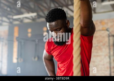 Uomo afro-americano appoggiato sulla corda da arrampicata vicino agli anelli della palestra, telaio della piattaforma all'interno della palestra, pausa allenamento Foto Stock