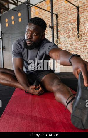 Uomo afroamericano che allunga la gamba su un tappeto rosso in palestra indossando lo smartwatch e usando anelli Foto Stock