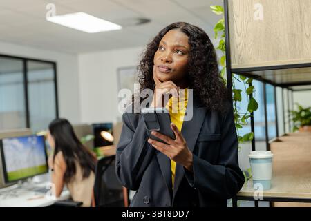 Diverse lavoratrici che indossano abiti che discutono idee in ufficio utilizzando smartphone e computer Foto Stock