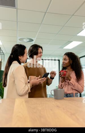 Diversi colleghi in piedi intorno al piano di lavoro in legno nella sala colazione dell'ufficio che reggono tazze e smartphone Foto Stock