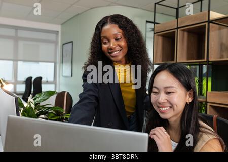 Diverse collaboratrici che collaborano con un computer portatile alla scrivania dell'ufficio con piante, oggetti d'arte, scaffali, sedie Foto Stock