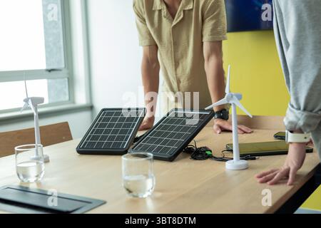 Colleghi asiatici che si appoggiano al tavolo in ufficio esaminando i pannelli solari pieghevoli e i modelli di turbine eoliche Foto Stock