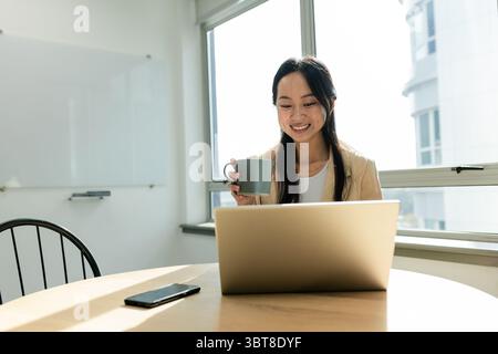 Donna asiatica che tiene la tazza da caffè e sorride al computer portatile sul tavolo dell'ufficio con smartphone e lavagna bianca Foto Stock