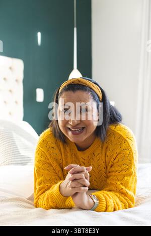 Donna sdraiata sullo stomaco a letto in camera da letto con cuscini testurizzati che indossano un maglione giallo e un cerchietto Foto Stock