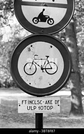 Cartello di divieto di bicicletta con il cartello inferiore: "FT INCLUSC - AKE DF ULP 10TOP", Whizgle News, Dutch Desk, Paesi Bassi, 1950 - 2000. Questi argomenti vengono visualizzati nell'immagine. Nella scena, ci sono due segni circolari montati su un palo. Il cartello superiore presenta una grafica di una motocicletta, resa semplice e stilizzata su sfondo bianco. Sotto di essa, il secondo cartello raffigura una bicicletta, anch'essa in stile minimalista, circondata da un bordo circolare. Lo sfondo mostra un ambiente naturale, probabilmente un parco, con note di verde visibili, che suggeriscono alberi nelle vicinanze. I colori nei segni presentano un st Foto Stock