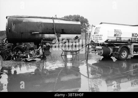 Resti di fuoco alla stazione di rifornimento Lijnden Lijnden, Whizgle News, Dutch Desk, Paesi Bassi, 1950 - 2000 il 29-06-1992. Questi argomenti sono mostrati nell'immagine. La scena cattura un ambiente industriale duro, dominato da grandi veicoli coinvolti nel trasporto di carburante. Un importante carro armato si trova sulla destra, con un logo aziendale distintivo in rosso e bianco brillante. È collegato tramite un tubo flessibile nero a un serbatoio cilindrico sulla sinistra, che appare più vecchio e più resistente agli agenti atmosferici, indicando anni di utilizzo. Il terreno è ricoperto da una lucentezza di acqua piovana, che riflette il cielo grigio sopra Foto Stock