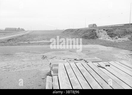 Piste per ingressi e uscite a Rijksweg costruzione di 6 strade messa a terra di strade, Whizgle News, Dutch Desk, Paesi Bassi, 1950 - 2000 su 28-11-1977. Questi argomenti sono mostrati nell'immagine. La scena raffigura un paesaggio spazioso e desolato caratterizzato da una tavolozza di colori tenui, dominata da sfumature di grigio. Una piattaforma di legno si trova in primo piano, composta da diverse tavole irregolari che appaiono intemprate, suggerendo un'esposizione prolungata agli elementi. A sinistra, un tratto di strada sterrata conduce in lontananza, dove curva leggermente, creando un senso di profondità. Parallelo alla r Foto Stock