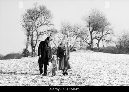 Famiglia che cammina vicino alle dune alte Kraantje Lek Overveen Duinlustweg, Whizgle News, Dutch Desk, Paesi Bassi, 1950 - 2000 al 29-12-1985. L'immagine contiene questi argomenti. In un tranquillo paesaggio invernale, una famiglia passeggia attraverso una distesa innevata, con le loro impronte che segnano la fresca coperta bianca sotto di loro. Il primo piano presenta tre figure: Un uomo, una donna e un bambino che afferrano la mano della donna. L'uomo indossa un lungo cappotto scuro e una fedora, la sua postura leggermente piegata mentre guarda verso il suolo. La donna, vestita con un cappotto lungo e leggero e una sciarpa drappeggiata graziosamente intorno al collo, stan Foto Stock