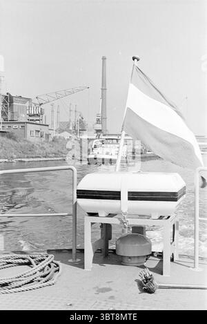 Gita in barca con Police Boat IJmuiden Police, Whizgle News, Dutch Desk, Paesi Bassi, 1950 - 2000 on 29-07-1975. Questi sono gli argomenti nell'immagine. La scena cattura una vista dei corsi d'acqua con un senso di attività industriale. In primo piano, una boa bianca e nera poggia su una nave, la sua posizione è fissata da una corda visibile. Dietro la boa, una bandiera, possibilmente nei colori del rosso e del bianco, fluttua dolcemente nella brezza, aggiungendo un vibrante contrasto con i toni monocromatici degli elementi circostanti. Diverse strutture industriali sono disposte lungo il fiume, con grandi fabbriche e gru che Foto Stock