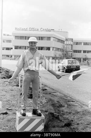Bambole come controllori del traffico Hoofddorp Paesi Bassi, Whizgle News, Dutch Desk, Paesi Bassi, 1950 - 2000 il 21-04-1991. Questi sono gli elementi dell'immagine. La scena presenta un manichino a grandezza naturale vestito con un giubbotto di sicurezza e un elmetto, in piedi su una piccola piattaforma posta ai margini di una carreggiata. Il manichino sembra dirigere il traffico, con le braccia allungate, uno con una piccola bandiera che presenta un'audace striscia rossa e bianca. Il vestito è giallo brillante, in netto contrasto con le sfumature monocromatiche dell'ambiente circostante. Dietro il manichino, c'è un mese Foto Stock
