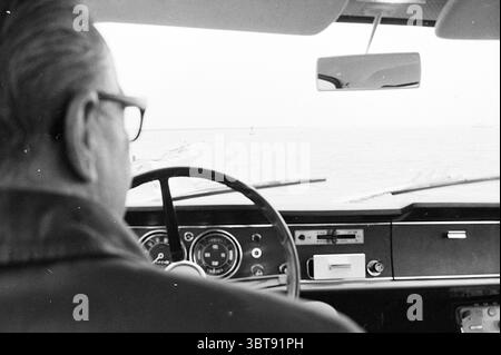 Tempesta sui moli e la costruzione del molo, Whizgle News, Dutch Desk, Paesi Bassi, 1950 - 2000 il 04-11-1969. L'immagine contiene questi argomenti. La scena cattura gli interni di un'auto d'epoca, concentrandosi sul conducente da una prospettiva retrovisiva. Il conducente, un uomo più anziano, viene visto con il suo profilo laterale evidenziato, mostrando capelli lisci sulla schiena e indossando occhiali dalla struttura spessa. La sua espressione trasmette concentrazione e forse nostalgia. Di fronte a lui, il volante è prominente, caratterizzato da un design classico con un mozzo centrale lucido e numerosi pulsanti e strumenti visibili sul cruscotto Foto Stock
