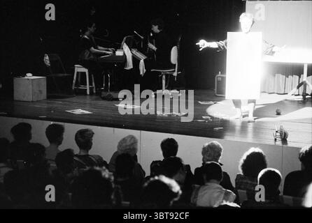 Sala concerti per la Festa della donna Women, Whizgle News, Dutch Desk, Paesi Bassi, 1950 - 2000 il 08-03-1990. Questi sono gli argomenti nell'immagine. La scena cattura uno spettacolo teatrale che si svolge su un palcoscenico poco illuminato. Al centro, un artista si erge con le braccia allungate, vestito con un abito bianco intenso che contrasta nettamente con l'oscurità circostante. La luce brillante dall'alto illumina la loro figura, creando un punto focale suggestivo in mezzo allo sfondo ombroso. A sinistra è visibile un piccolo ensemble, con un pianista seduto su una tastiera e un altro musicista posizionato nelle vicinanze, entrambi Foto Stock