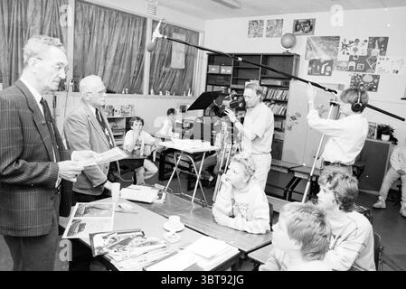 Registrazione TV Beatrixschool Zandvoort Zandvoort, Whizgle News, Dutch Desk, Paesi Bassi, 1950 - 2000 il 17-04-1991. Questi sono gli elementi dell'immagine. In una luminosa aula, due uomini sono in prima linea, impegnati in una conversazione. Un uomo, vestito con un abito scuro e un paio di occhiali, sembra spiegare qualcosa con passione, mentre l'altro, con un abito più leggero, ascolta con attenzione. Entrambi trasudano un senso di autorità e di conoscenza. Dietro di loro, un piccolo gruppo di studenti siede alle scrivanie di legno, attento e impegnato. Le loro espressioni vanno dalla curiosità alla concentrazione, alto Foto Stock
