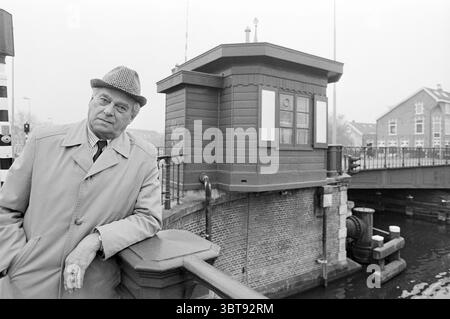 Sig. Deen WWII raid a Haarlem Haarlem, Paesi Bassi, Whizgle News, Dutch Desk, Paesi Bassi, 1950 - 2000 il 30-11-1994. L'immagine include questi argomenti. In questa scena, un uomo si trova accanto ad un canale, appoggiato contro una ringhiera. Indossa un trench di colore chiaro e un cappello a fantasia che aggiunge un tocco di carattere. La sua espressione è seria, suggerendo contemplazione o nostalgia. Lo sfondo presenta una piccola struttura in legno, possibilmente una sala di controllo o una cabina, con grandi finestre e un tetto piatto. L'acqua del canale è calma, riflettendo i colori tenui dell'ambiente circostante. C'è un brid Foto Stock