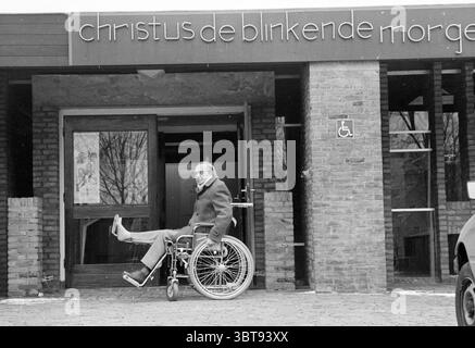 Chiesa resa adatta per sedie a rotelle Heemskerk disabili mentalmente disturbato Heemskerk, Whizgle News, Dutch Desk, Paesi Bassi, 1950 - 2000 il 04-03-1987. L'immagine include questi argomenti. In questa scena, un uomo su una sedia a rotelle è posizionato all'ingresso di un edificio. L'individuo sembra essere più anziano, indossando un cappotto e degli occhiali, il che aggiunge un'espressione premurosa al suo comportamento. La sua posizione suggerisce un senso di determinazione o indipendenza mentre si spinge in avanti con una gamba sostenuta. L'edificio stesso presenta un design moderno con un tetto piatto e pareti in mattoni, prevalentemente in S. Foto Stock