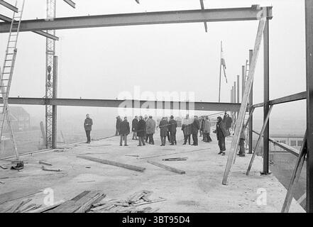 Un gruppo di persone guarda il guscio d'acciaio di un edificio di fabbrica, Whizgle News, Dutch Desk, Paesi Bassi, 1950 - 2000. Questi argomenti sono mostrati nell'immagine. In un ambiente industriale nebbioso, un gruppo di persone si riunisce in un cantiere di scheletri. L'atmosfera è attenuata, con una tavolozza di grigi che domina la scena. Le travi in acciaio si sollevano verso l'alto nella foschia, creando una struttura lineare astratta contro il cielo opaco. Diversi individui si trovano in agglomerati, rivestiti di cappotti scuri, dando un senso di serietà all'umore. Le loro posture suggeriscono un mix di contemplazione e discussione, mentre ap Foto Stock