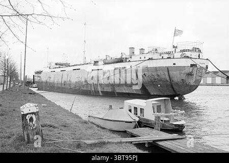 Nave Maro Havenstr. Zaandam Ships Zaandam Havenstraat, Whizgle News, Dutch Desk, Paesi Bassi, 1950 - 2000 il 02-04-1977. L'immagine contiene questi argomenti. La scena presenta una grande nave cargo ormeggiata lungo il bordo di un fiume o di un canale. La nave, in particolare invecchiata, mostra uno scafo arrugginito con vernice che peeling, che mostra sfumature di grigio e accenni di toni più scuri, riflettendo la sua lunga esposizione agli elementi. La posizione ancorata crea un punto focale forte, dominando il paesaggio. In primo piano, una stretta tavola di legno conduce dalla riva alla nave, suggerendo l'accesso per i lavoratori o V Foto Stock