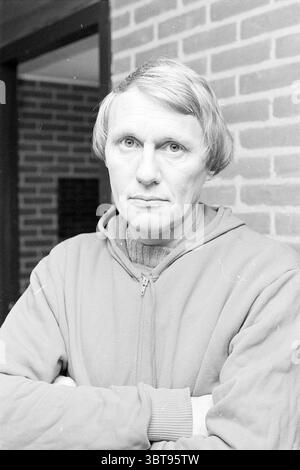 Portraits Jan Visscher V.V.B. Portraits, Whizgle News, Dutch Desk, Paesi Bassi, 1950 - 2000 il 12-11-1976. L'immagine mostra questi argomenti. In questa scena, un uomo sta con le braccia incrociate, appoggiandosi leggermente contro quello che sembra essere un muro testurizzato di mattoni marrone-rossastro. Ha capelli ondulati e di colore chiaro che incornicia il suo viso, che porta un'espressione seria. I suoi occhi, di colore chiaro e leggermente ristretti, emanano un senso di introspezione o contemplazione. L'uomo è vestito con una morbida felpa con cappuccio e zip, che è una tenue tonalità, forse grigio chiaro o beige, che contrasta con il calore del t Foto Stock