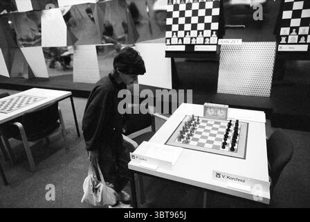 Blastovens Chess Tournament Wijk aan Zee, Whizgle News, Dutch Desk, Paesi Bassi, 1950 - 2000 il 19-01-1993. Questi sono gli elementi dell'immagine. La scena raffigura un ambiente di torneo di scacchi, dominato da una paletta in bianco e nero. Un giovane, probabilmente concentrato e contemplativo, si sta piegando leggermente in avanti, esaminando una scacchiera di fronte a loro. Il tavolo è quadrato e presenta una configurazione standard degli scacchi, con i pezzi disposti nelle loro posizioni iniziali. Sullo sfondo, una serie di grandi scacchiere viene visualizzata su cavalletti o pareti, mostrando varie posizioni da matche multiple Foto Stock