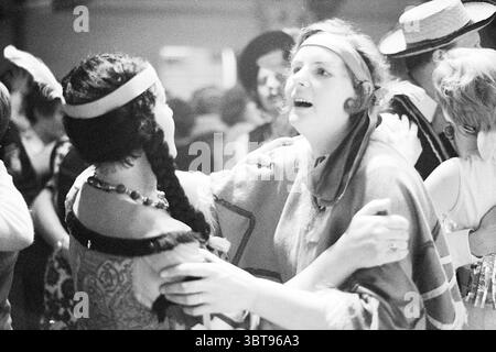 Carnevale per anziani Noordwijkerhout Carnevale, Whizgle News, Dutch Desk, Paesi Bassi, 1950 - 2000 il 10-02-1970. L'immagine contiene questi argomenti. La scena cattura un'atmosfera vivace, probabilmente da una festa vintage o da un evento di danza. In primo piano, due donne sono impegnate in un abbraccio animato, le loro espressioni piene di gioia ed eccitazione. Uno ha lunghi capelli scuri intrecciati, ed è adornata da una collana con perline che aggiunge un tocco di lucentezza al suo outfit a motivi geometrici, che evoca un senso di stile storico. La seconda donna, con i capelli ricci più leggeri, indossa un indumento fluente Foto Stock