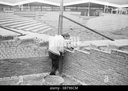 Edificio punto di discussione Zeewijkplein IJmuiden costruzione pozzi IJmuiden Zeewijkplein Paesi Bassi, Whizgle News, Dutch Desk, Paesi Bassi, 1950 - 2000 il 21-02-1972. Questi sono gli elementi dell'immagine. La scena raffigura un cantiere immerso in una tavolozza monocromatica, con sfumature di grigio che dominano il paesaggio visivo. In primo piano, una persona, vestita con un maglione di colore chiaro e pantaloni scuri, viene piegata, lavorando intensamente su un muro di mattoni in costruzione. La consistenza dei mattoni esposti è prominente, mostrando la finitura ruvida e la sottile varietà Foto Stock