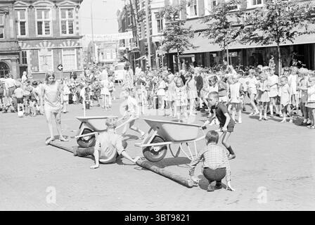 Apertura dei giochi per le vacanze giochi per le vacanze vacanze vacanze, Whizgle News, Dutch Desk, Paesi Bassi, 1950 - 2000 il 01-07-1969. Questi sono gli argomenti nell'immagine. La scena cattura una strada movimentata piena di attività ed energia. In primo piano, due bambini sono impegnati in una giocosa competizione utilizzando carriole, ognuna posizionata in cima a un tronco che funge da trave di bilanciamento improvvisata. Le loro espressioni riflettono la concentrazione e l'entusiasmo mentre affrontano la sfida. Intorno a loro, un gruppo eterogeneo di bambini e pochi adulti si trovano in un semicerchio, osservando con anticipo. Foto Stock