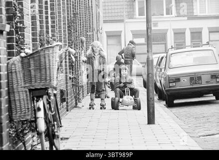Quartiere in vista: Indische Buurt Zuid Haarlem Paesi Bassi, Whizgle News, Dutch Desk, Paesi Bassi, 1950 - 2000 il 17-03-1980. L'immagine contiene questi argomenti. La scena cattura una strada vivace di un'epoca passata, evocando un senso di nostalgia. In primo piano ci sono due bambini: Una ragazza sui pattini a rotelle, che indossa un caldo cappotto rivestito di pelliccia che contrasta con lo sfondo mite, e un ragazzo seduto in un piccolo giocattolo colorato che ricorda un go-kart. La sua espressione giocosa è visibile, trasmettendo un senso di gioia e avventura. La strada e' fiancheggiata da edifici in mattoni, le loro facciate mostrano vari elementi Foto Stock
