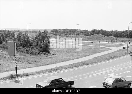 Panoramica del terreno Langevelderslag Noordwijkerhout terrains Noordwijkerhout Langevelderslag, Whizgle News, Dutch Desk, Paesi Bassi, 1950 - 2000 il 05-07-1977. L'immagine contiene questi argomenti. La scena raffigura un incrocio stradale con un sentiero tortuoso che porta in lontananza, fiancheggiato da una vegetazione lussureggiante su un lato. Il terreno è prevalentemente erboso, evidenziando uno spazio naturale e aperto che contrasta con l'asfalto della strada. Si possono vedere due veicoli: Un'auto classica di colore scuro che si muove verso destra e un'altra auto più piccola che si avvicina da sinistra. Le auto aggiungono un senso di movimento in mezzo Foto Stock