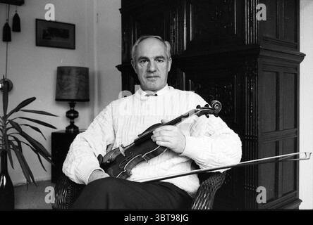 Violinista Gijs Beths S'poort Santpoort, Whizgle News, Dutch Desk, Paesi Bassi, 1950 - 2000 il 17-02-1991. L'immagine contiene questi argomenti. La scena presenta un uomo seduto in uno spazio interno accogliente e delicatamente illuminato. È vestito con un comodo maglione a maglia che si distingue per le tonalità scure dei mobili di fondo. La sua espressione è calma e premurosa, mentre tiene un violino in una mano, appoggiandolo contro le sue ginocchia, mentre l'arco giace sull'altro braccio. Intorno a lui c'è un ambiente invitante. Sullo sfondo, un grande pezzo di legno, possibilmente un mobile ornato, aggiunge un tocco Foto Stock