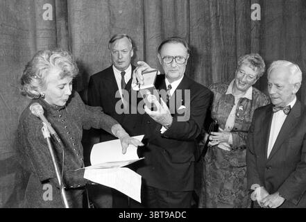 Medaglia onoraria Alberdingk Thijm associazione teatrale H'kerk Heemskerk, Whizgle News, Dutch Desk, Paesi Bassi, 1950 - 2000 il 10-11-1991. Questi argomenti vengono visualizzati nell'immagine. In un ambiente scarsamente illuminato, un gruppo di quattro individui è riunito, creando un senso di attenzione su un momento di significato. Lo sfondo è una tenda dalle texture morbide che dona un'atmosfera formale all'occasione con i suoi toni tenui. A sinistra, una donna con un indumento scuro e testurizzato si piega leggermente in avanti, concentrandosi sulle carte nelle sue mani. I suoi capelli sono modellati con onde morbide, e trasuda un senso di portamento e importanza A. Foto Stock