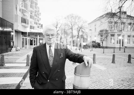 Sig. Davids nuovo presidente KNOV Associations, Whizgle News, Dutch Desk, Paesi Bassi, 1950 - 2000 il 10-01-1990. L'immagine contiene questi argomenti. In un vivace ambiente urbano, un uomo di mezza età si erge con sicurezza su un passaggio pedonale pavimentato. È vestito con un abito scuro, i revers ben definiti, con una cravatta a motivi geometrici che aggiunge un tocco di raffinatezza. L'uomo ha i capelli ben disegnati, incorniciati da un paio di occhiali che enfatizzano il suo comportamento. Intorno a lui ci sono elementi di una vivace scena di strada. A sinistra, un edificio moderno con una facciata in vetro contrasta con il più antico, più tradizionale archite Foto Stock