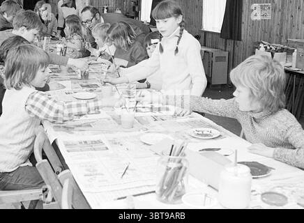 Klieder kladder klodder party Noordervreugd Children's Homes Children's Party Children's Party Children, Whizgle News, Dutch Desk, Paesi Bassi, 1950 - 2000 il 17-04-1973. L'immagine contiene questi argomenti. In un ambiente interno vivace, un lungo tavolo è pieno di bambini impegnati in un'attività creativa, probabilmente dipingendo. La camera presenta un rivestimento rustico in legno che le dona un'atmosfera calda e invitante. La luce del sole scorre attraverso grandi finestre, illuminando la scena e proiettando ombre morbide. I bambini, vestiti con un mix di maglioni colorati e abiti a fantasia, sono concentrati sui loro compiti. Alcuni sono "ho" Foto Stock