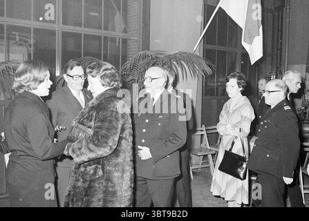 Addio Burg. De Gou di H'lem vigili del fuoco Sindaci dei vigili del fuoco, Whizgle News, Dutch Desk, Paesi Bassi, 1950 - 2000 il 16-12-1976. Questi sono gli elementi dell'immagine. La scena raffigura un raduno formale, ambientato all'interno di uno spazio interno caratterizzato da architettura industriale, tra cui grandi finestre che consentono alla luce ambientale di filtrare. L'atmosfera è seria ma cordiale, evidente nelle espressioni e nel linguaggio del corpo degli individui presenti. In primo piano, due donne si impegnano in conversazione. Una donna, vestita con un abito scuro ed elegante, ha i capelli a spalla ed è apparsa Foto Stock