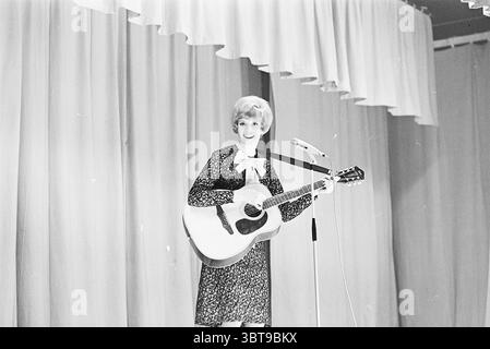 Ria Valk con chitarra Andre Meurs Cabaret, Whizgle News, Dutch Desk, Paesi Bassi, 1950 - 2000 il 13-04-1966. L'immagine contiene questi argomenti. In un ambiente scenico, un esecutore è in piedi con sicurezza con una chitarra acustica in mano. È vestita con un abito scuro a motivi geometrici caratterizzato da un delicato design floreale, completato da un arco oversize bianco intorno al collo. La sua acconciatura è elegantemente composta da onde morbide, che inquadrano il suo viso e aggiungono un tocco di fascino al suo aspetto. Lo sfondo è costituito da tessuto drappeggiato in colori tenui e tenui, che creano un'atmosfera liscia ed elegante. Le tende hanno una gent Foto Stock