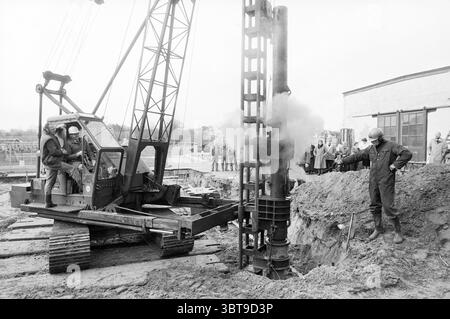 Per costruire. Alla guida del primo mucchio., Whizgle News, Dutch Desk, Paesi Bassi, 1950 - 2000 dal 04-03-1988. Questi sono gli elementi dell'immagine. Sulla scena, un cantiere si dispiega con una combinazione di macchinari industriali e lavoratori impegnati nelle loro attività. A dominare il primo piano c'è un grande carro di perforazione, la cui struttura è composta da metallo robusto e verniciata in un colore tenue, possibilmente verde o nero, che mostra segni di usura e utilizzo. Il carro è alto, con un albero alto e varie leve, a indicare la sua complessità operativa. A sinistra, due lavoratori sono seduti all'interno della macchina, rivestiti in sicurezza Foto Stock